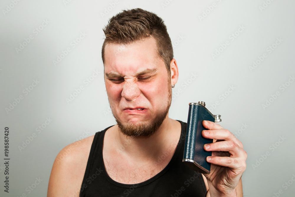 Young bearded guy drinks alcohol from a flask. On gray background. Young bearded guy drinks alcohol from a flask. On gray background.
