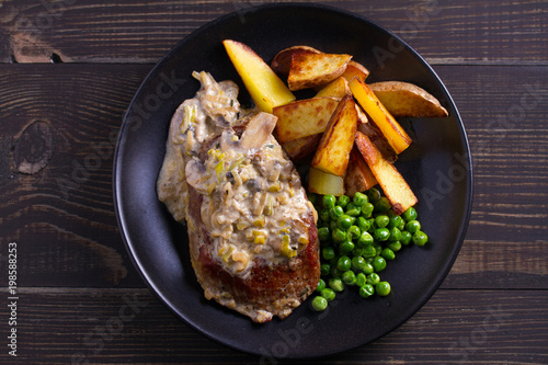 Beef steak diane with mushroom and leek cream sauce, potato fries and green peas in black plate on wooden background. overhead