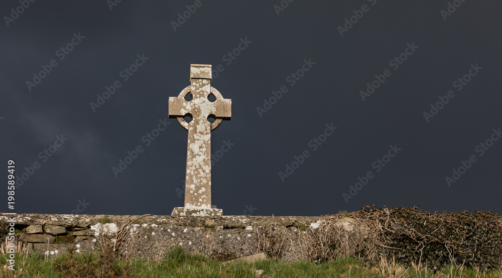 Celtic cross gravestone behind soon wall of a cemetery in rural Ireland, dark stormy rain clouds and sunlight Celtic cross gravestone behind soon wall of a cemetery in rural Ireland, dark stormy rain clouds and sunlight