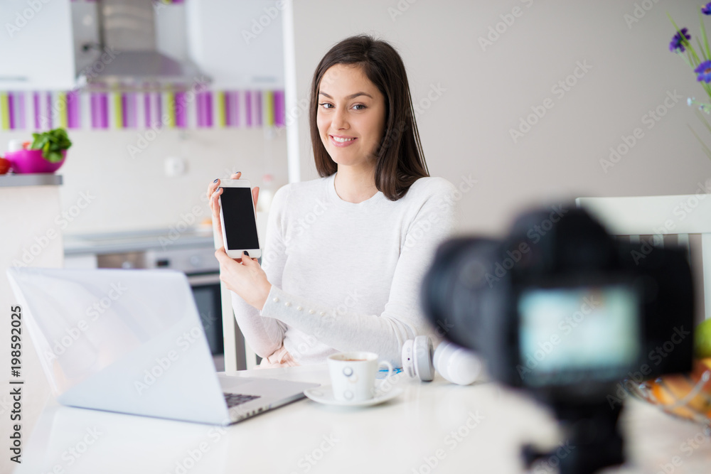Young happy beautiful girl is sitting at the kitchen table and showing the phone to the camera with laptop in front of her while drinking coffee. Young happy beautiful girl is sitting at the kitchen table and showing the phone to the camera with laptop in front of her while drinking coffee.