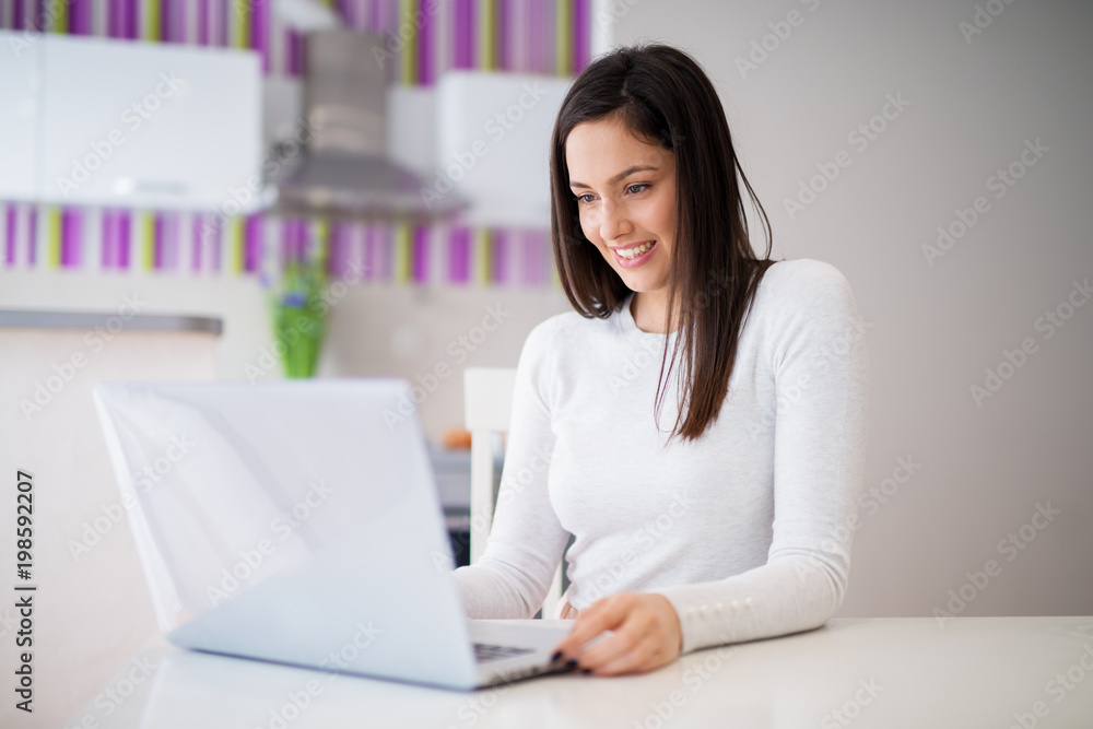 Young beautiful cheerful girl is sitting at the kitchen table and is happy with what she sees on her laptop. Young beautiful cheerful girl is sitting at the kitchen table and is happy with what she sees on her laptop.