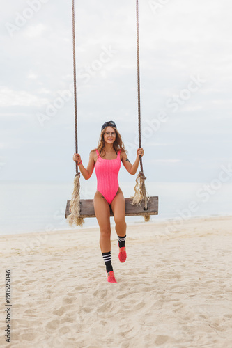 young woman in pink swimsuit sitting on swing on beach at resort