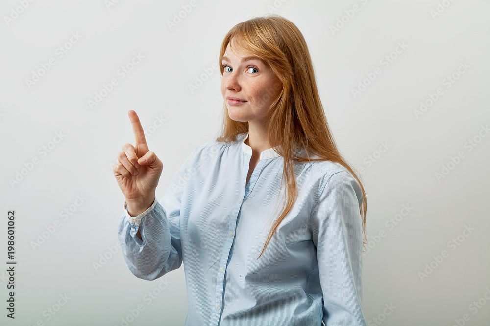 Headshot portrait of young european lady with red hair, white skin and freckles looking at the camera and showing at the information on left side of the picutre with her index finger raised.