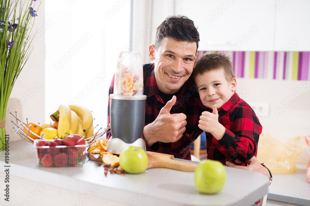 Father and son in same shirt are giving a thumbs up together while waiting for a blender to finish making their smoothies.