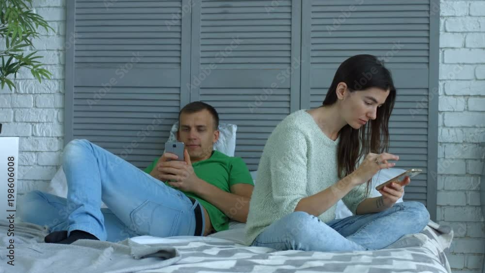 Attractive couple phubbing, busy with personal devices and networking while spending free time in bedroom. Handsome man and woman addicted to smart phones and games online ignoring each other at home. Attractive couple phubbing, busy with personal devices and networking while spending free time in bedroom. Handsome man and woman addicted to smart phones and games online ignoring each other at home.
