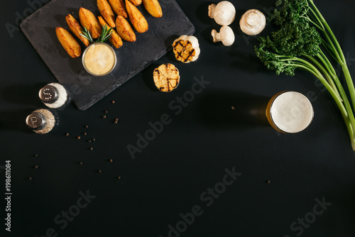 top view of delicious baked potatoes with sauce, spices and glass of beer on black