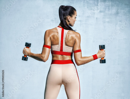 Athletic woman doing exercise for arms. Photo of muscular fitness model working out with dumbbells on grey background. © Romario Ien