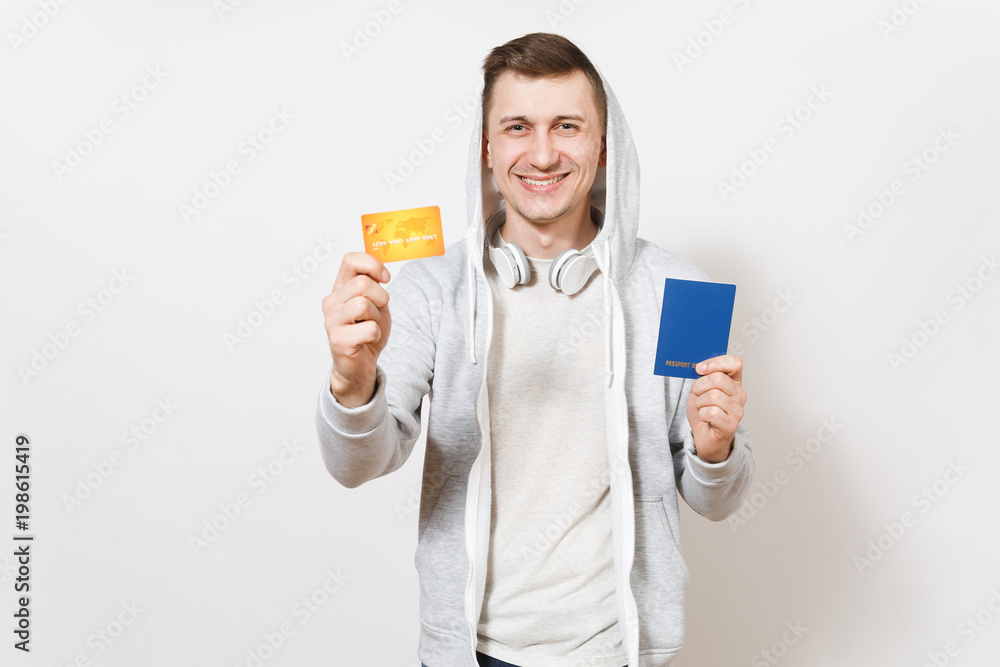 Young handsome smiling happy man in t-shirt, light sweatshirt with hood with headphones holds international passport and credit card in hands isolated on white background. Concept of travel, tourism Young handsome smiling happy man in t-shirt, light sweatshirt with hood with headphones holds international passport and credit card in hands isolated on white background. Concept of travel, tourism