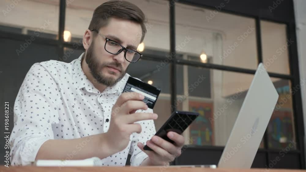 One young male person in glasses sit in workspace, hold cellphone and looking shop on line. Consumer pay by contactless debit bankcard. Commercial concept of buyer use the currency by telephone One young male person in glasses sit in workspace, hold cellphone and looking shop on line. Consumer pay by contactless debit bankcard. Commercial concept of buyer use the currency by telephone