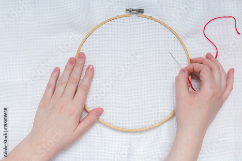 Hands of teenage girl with needle on embroidery frame on white textile background canveplian top view flat lay  photo