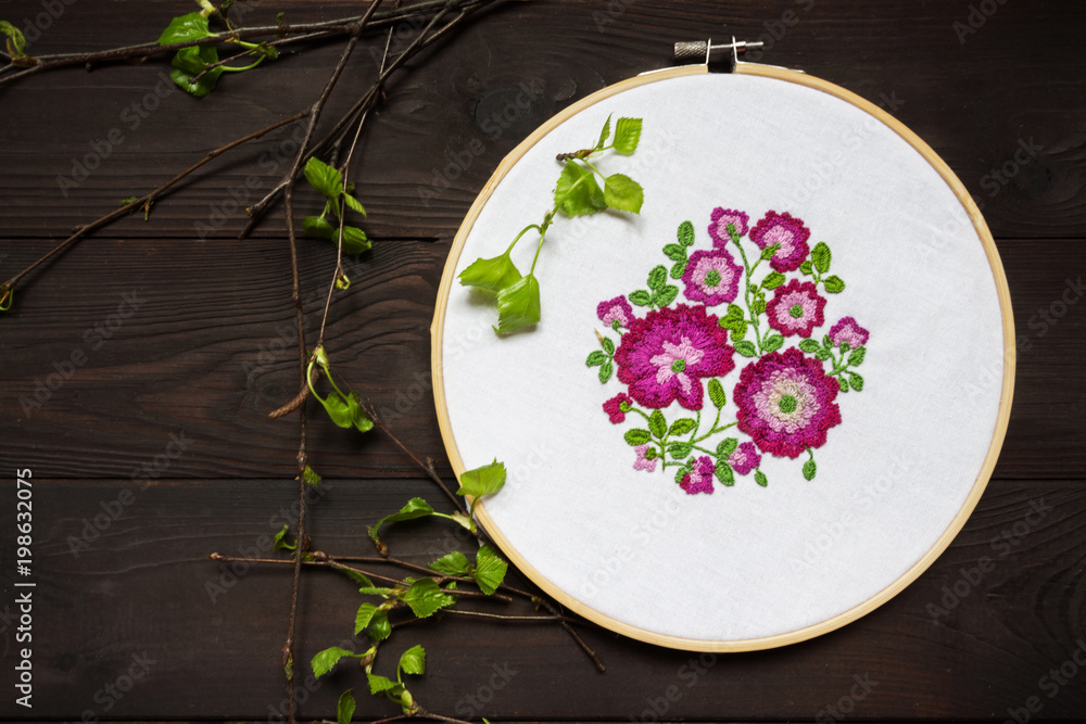 Embroidery of flowers on a white canvas in a frame on a dark wooden background in a rustic style with branches of trees with young leaves