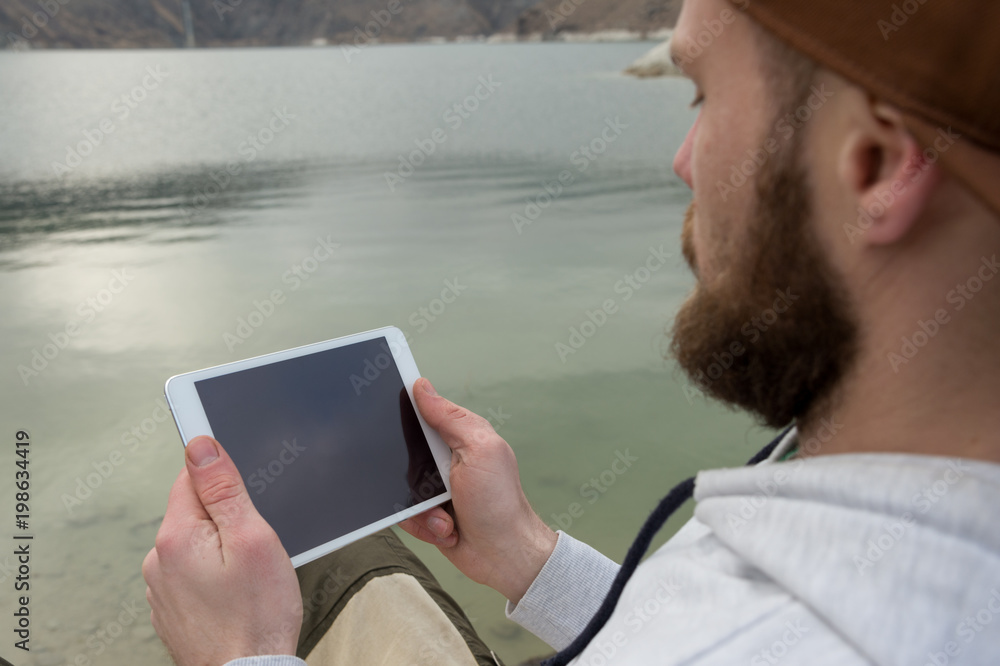 Close-up of a hipster in a brown cap in the open air holds a white tablet pc in his hands. A bearded man looks at the tablet. OTS view from behind the shoulder