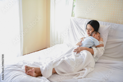 Newborn Concept. Mother and child on a white bed. Mom and baby boy playing in bedroom. Parent and little kid relaxing at home. Family having fun together. Newborn baby is fussing and crying.