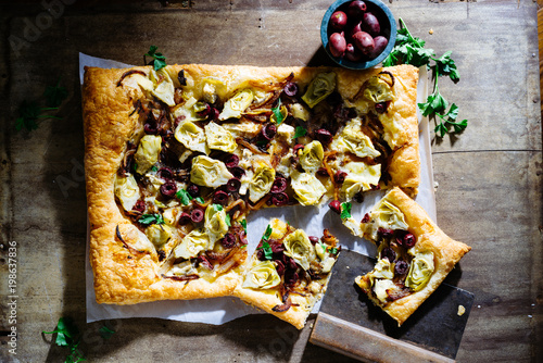 Foto Overhead of artichoke heart, black olive and caramelized onion tart on wooden surface with pieces cut out topped with parsley and small bowl of black olives