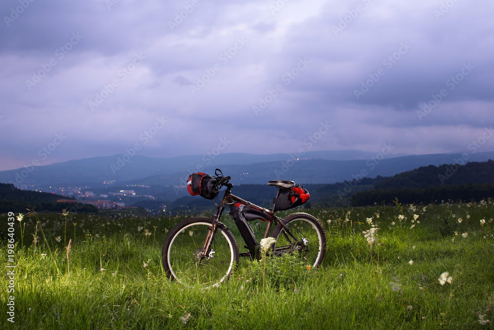 mountain bike after the bike ride and transport bags on a lush green grass at sunset amid the mountain peaks in the background