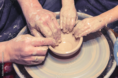 A close-up of a man potter teaches a child how to properly mold a bowl of brown clay on a potter's wheel in a bright workshop, a top view.