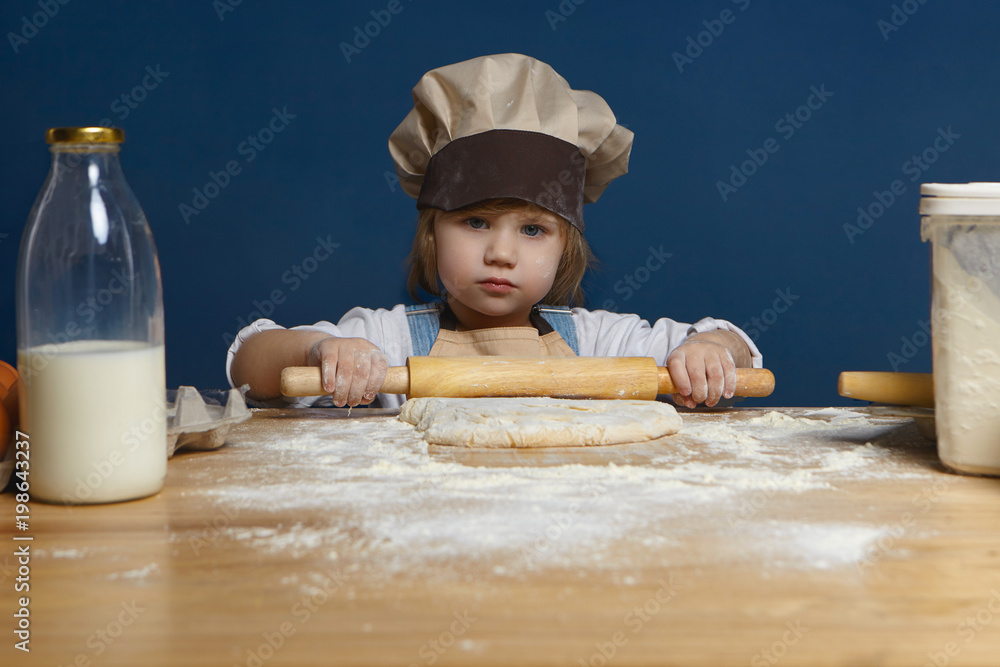 Portrait of serious cute female kid of preschool age standing at kitchen counter wearing chef hat, flattening dough using rolling pin, helping mother to bake apple pie. Cooking, food and bakery Portrait of serious cute female kid of preschool age standing at kitchen counter wearing chef hat, flattening dough using rolling pin, helping mother to bake apple pie. Cooking, food and bakery