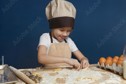 Adorable 8 year old male child in beige apron and hat standing in kitchen, molding cookies on counter, trying hard, sticking out tongue. Focused little boy making pasty at table covered with flour