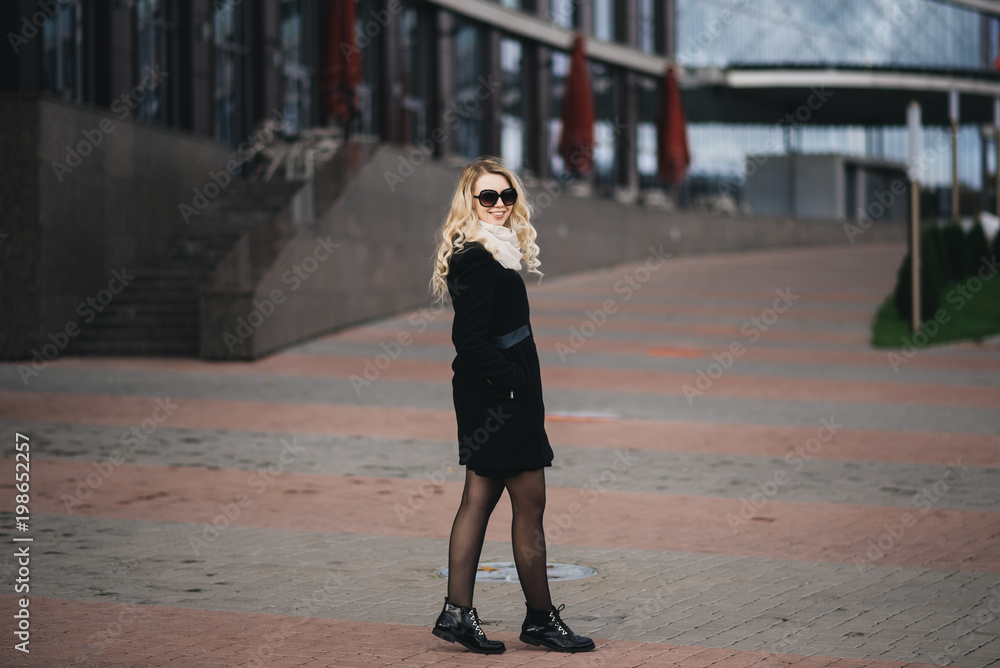 Beautiful young girl with blond wavy hair in a black coat against the backdrop of modern buildings