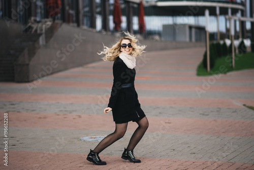Beautiful young girl with blond wavy hair in a black coat fun on background of modern building