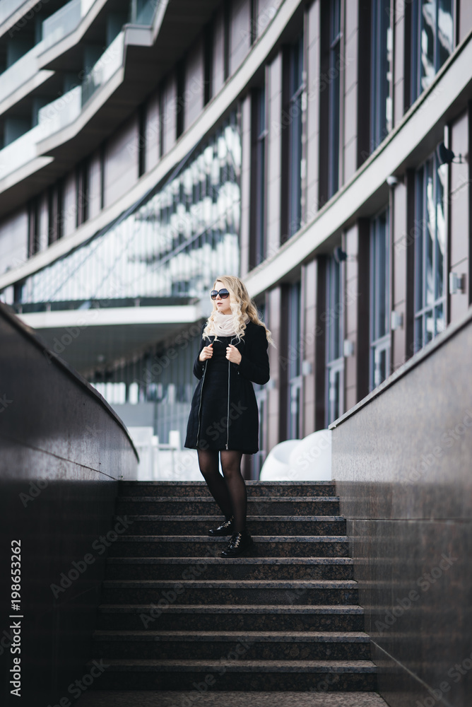 Beautiful young girl with blond wavy hair in black dress goes against the background of modern building
