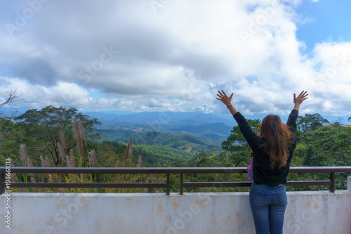 back view of asian happy female in casual outfit looking at amazing panoramic view of mountain and jungle. woman raise her hand feeling happiness or success.