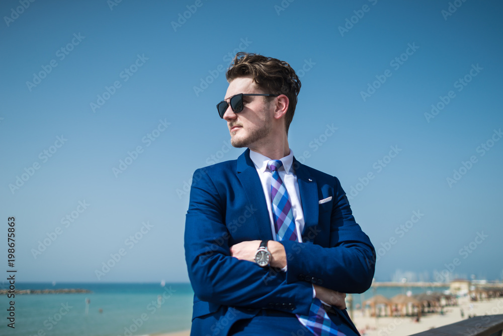 Young serious man in elegant suit and sunglass on the background of the sea and sky. Successful manager is relaxing on the nature in warm sunny day. Trendy guy stands on city promenade