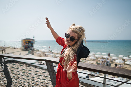 Young attractive woman in bright red dress dances on the city promenade. Elegant blonde girl active gesticulate on background of sea and sky. Funny female traveler has fun near beach