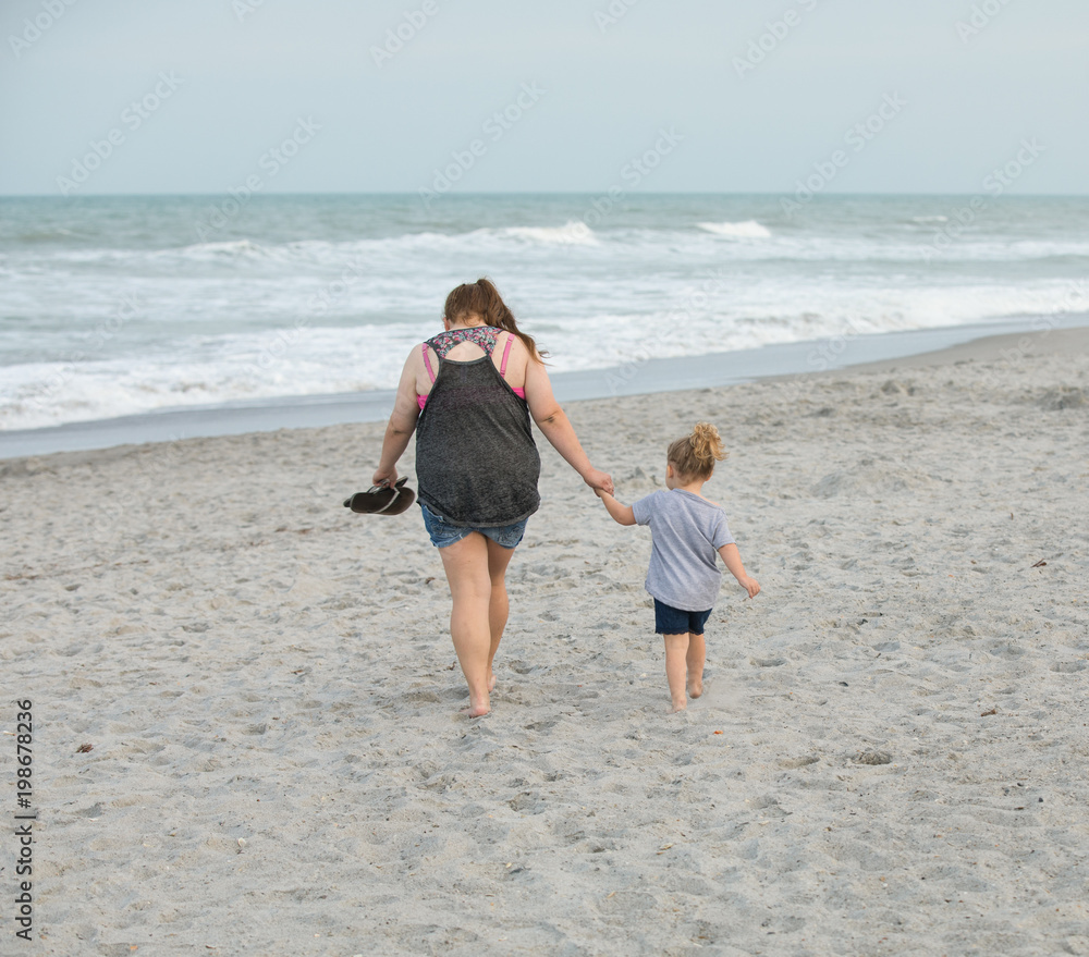 unknown mother and daughter hold hands while walking barefoot on the beach at sunset