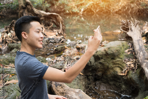 Selfie photo concept : Asian young man holding smartphone for selfies at  forest, beautiful scenery for peachful in spring , It shows moisture, tranquility and refreshing of rainforest in Thailand photo