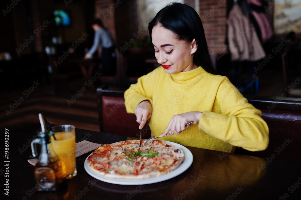 Funny brunette girl in yellow sweater eating pizza at restaurant.