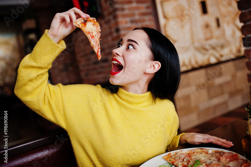 Funny brunette girl in yellow sweater eating pizza at restaurant.