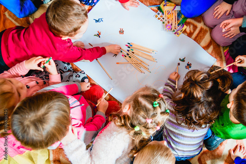 several small children draw on a sheet of paper with pencils. A view from above.