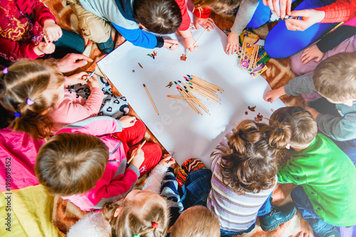 several small children draw on a sheet of paper with pencils. A view from above.
