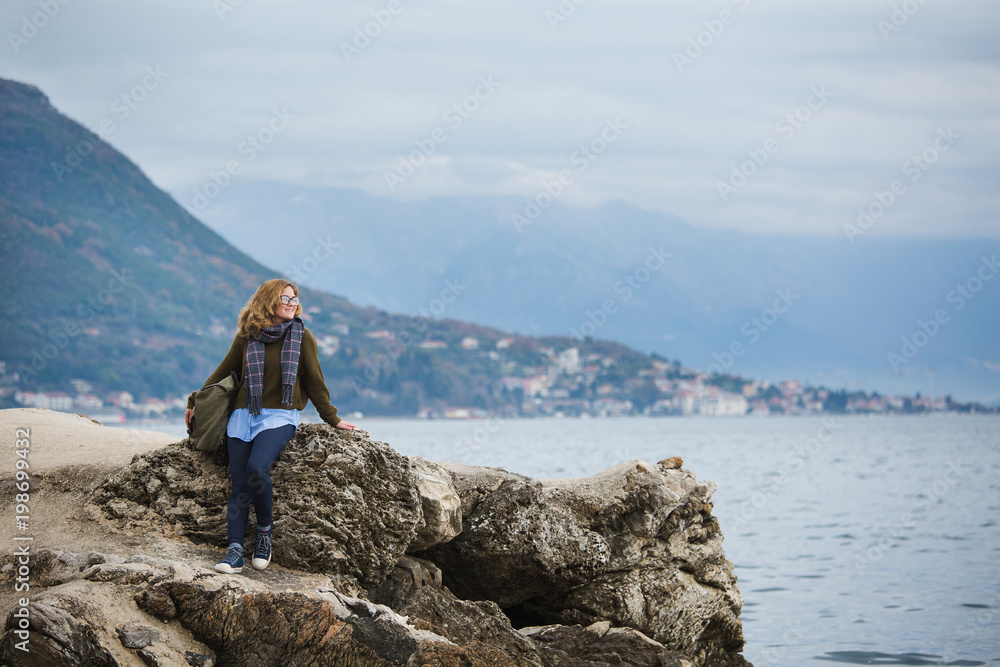 Woman traveler by the sea. On the horizon high mountains. Girl dressed in a sweater and scarf.