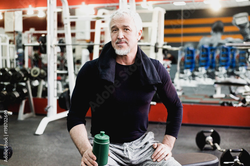 Adult man with gray hair resting after workout in gym with green bottle of water photo