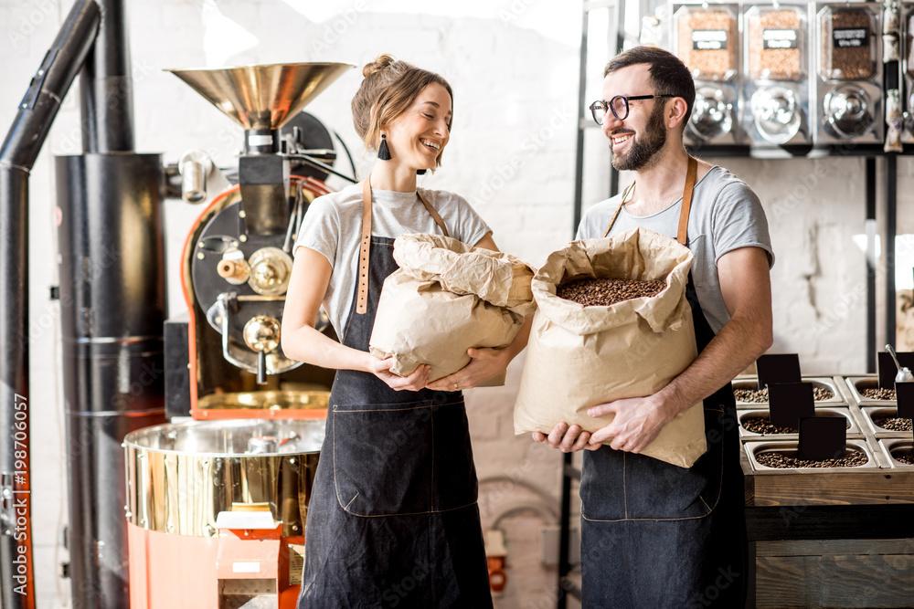 Portrait of a two happy baristas in uniform standing with bags full of coffee beans at the coffee store
