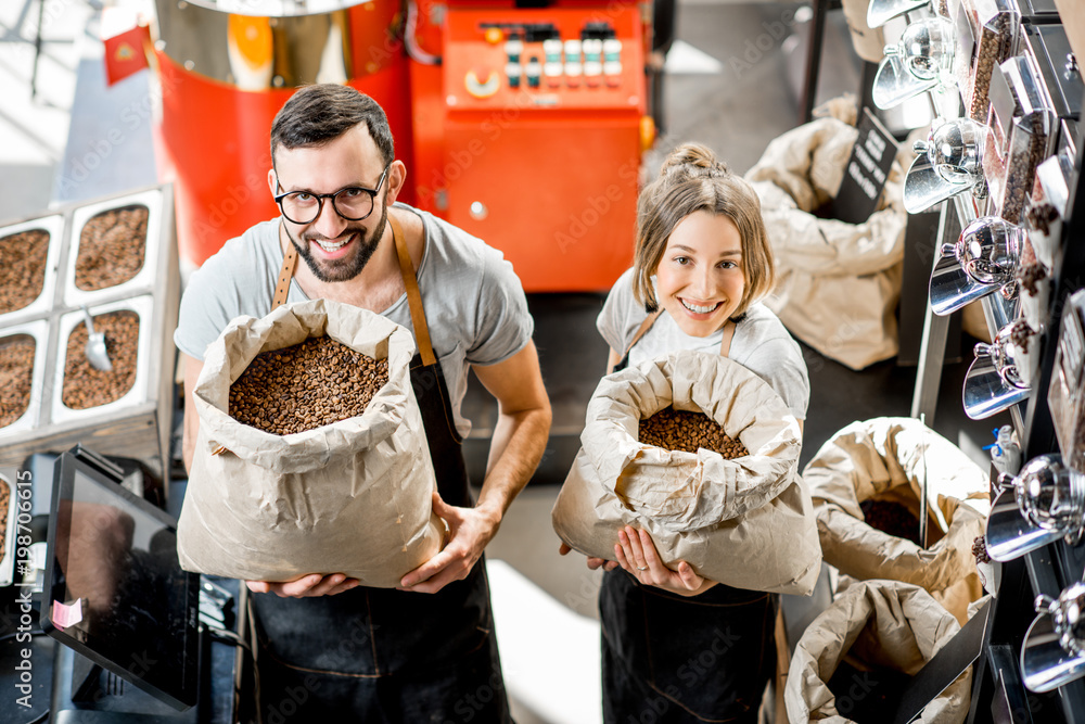 Portrait of a two happy sales persons in uniform standing with bags full of coffee beans in the coffee store