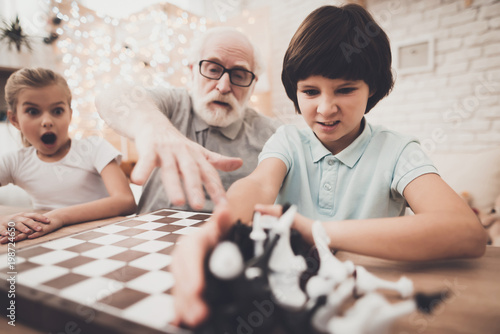 Grandfather, grandson and granddaughter at home. Grandpa teaches children how to play chess.