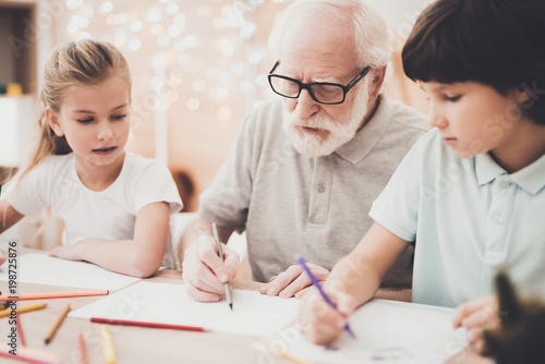 Grandfather, grandson and granddaughter at home. Children are drawing with color pencils.
