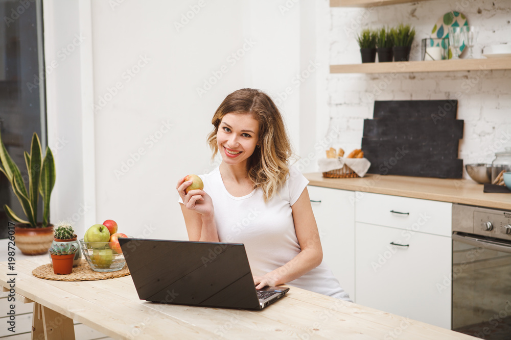 Attractive woman sitting at a kitchen table behind a laptop holding an apple in her hand looking into the camera and smiling. Work at home. Lifestyle. Space for text. Attractive woman sitting at a kitchen table behind a laptop holding an apple in her hand looking into the camera and smiling. Work at home. Lifestyle. Space for text.