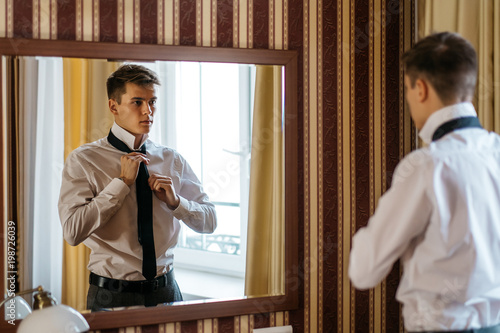 A young handsome man ties up a tie in front of a mirror