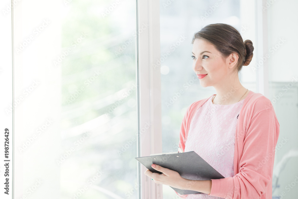 Business woman standing working near a window and holding clipboard at office. Business woman standing working near a window and holding clipboard at office.