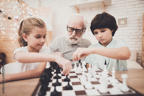 Grandfather, grandson and granddaughter at home. Children and grandpa are playing chess.