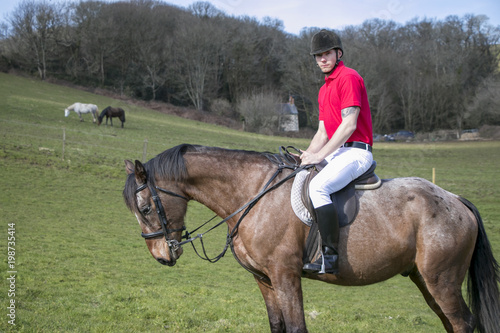 Rider on Horseback in field, wearing red polo shirt, white trousers, black boots with horses in the background