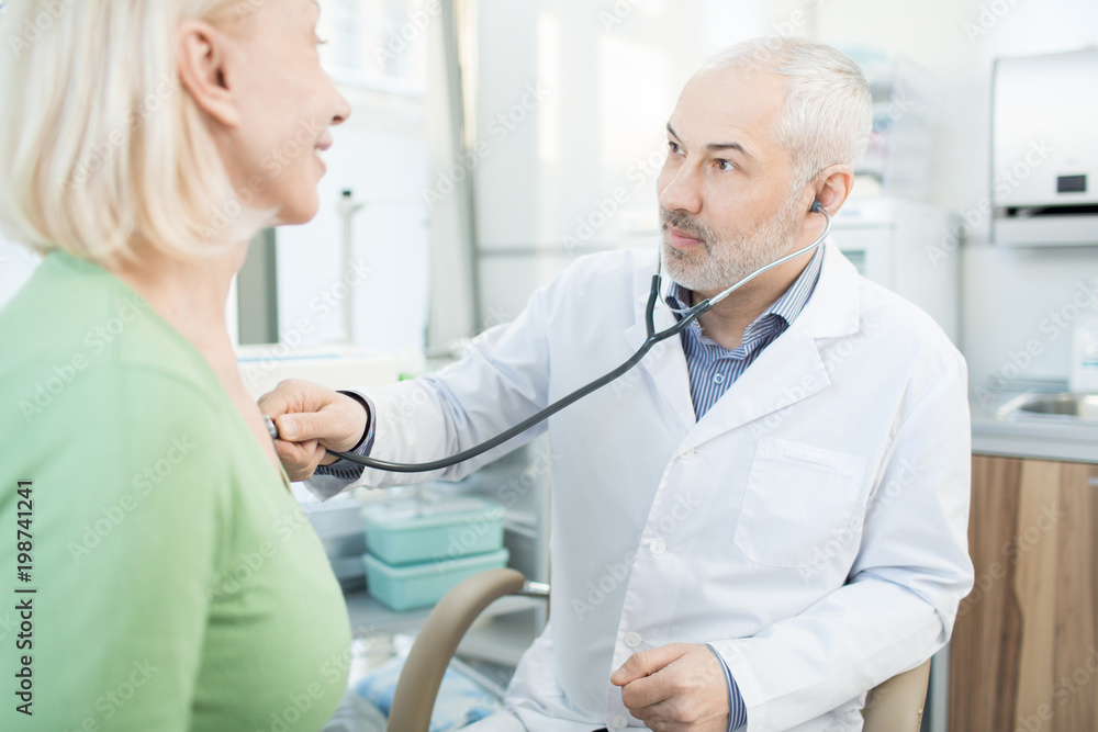 Obraz premium Mature doctor in whitecoat looking at his patient during treatment with stethoscope
