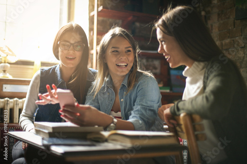 Three smiling students girls having conversation and using smart phone.