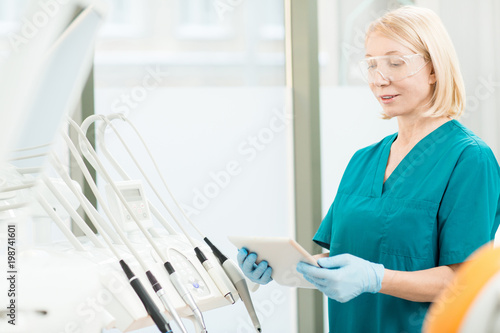 Mature professional dentist in protective eyeglasses, gloves and uniform using modern gadget to look for necessary information © pressmaster