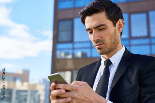 Young employee in suit and tie reading or writing message in smartphone in urban environment