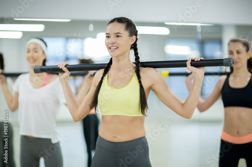 Young active woman and her groupmates with gymnastic bars on shoulders exercising in fitness center
